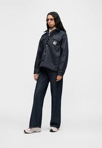 Σκούρο τζιν μπουφάν με χρυσαφένιες λεπτομέρειες και μπροστινές τσέπες, σετ με φαρδιά σκουρόχρωμα τζιν παντελόνια και ανοιχτόχρωμα αθλητικά παπούτσια. Το μοντέλο έχει μακριά, κυματιστά μαλλιά.