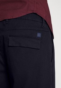 Nærbillede af marineblå teksturerede bukser med klaplomme og lille mærkeetiket, kombineret med en vinrød skjorteunderkant.