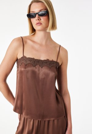 Kvinde iført brun satin camisole med blonder og matchende bukser, tilbehør med smalle sorte solbriller, stående mod hvid baggrund.