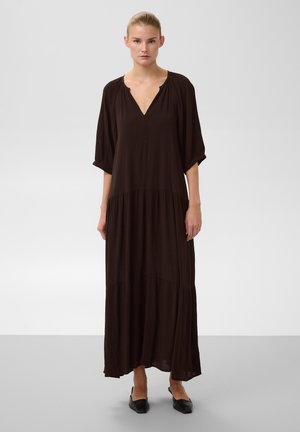 Femme debout portant une longue robe marron foncé à col en V avec des manches trois-quarts et des chaussures plates noires sur un fond uni.