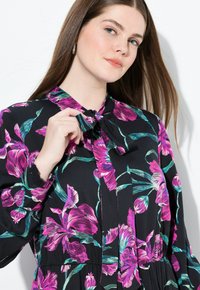 Ung kvinde med langt brunt hår iført en sort kjole med rosa og turkise blomstermønstre, der justerer båndet ved halsudskæringen.