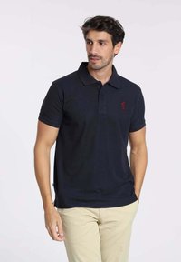 Polo shirt navy en coton doux avec un col classique, des manches courtes et un petit logo rouge sur la poitrine, associé à un pantalon beige.
