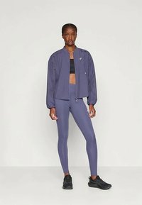 Moteris stovi atsipalaidavusi, vilkėdama purpurinį zip-up džemperį, atitinkančius legginus, juodą sportinį liemenėlę ir juodus sportinius batus.