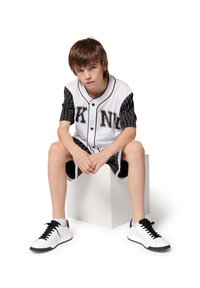 Tenue de base-ball pour jeunes avec un corps blanc, des manches à rayures noires, un short noir et des baskets blanches. Design simple et sportif.