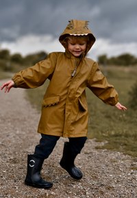 Brown waterproof raincoat with a hood featuring a dinosaur design, paired with black rubber boots, standing on a gravel path.