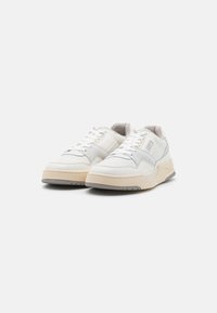 White sneakers featuring mesh and suede accents, rounded toe, thick ridged sole, and prominent stitching details. Minimalist design throughout.