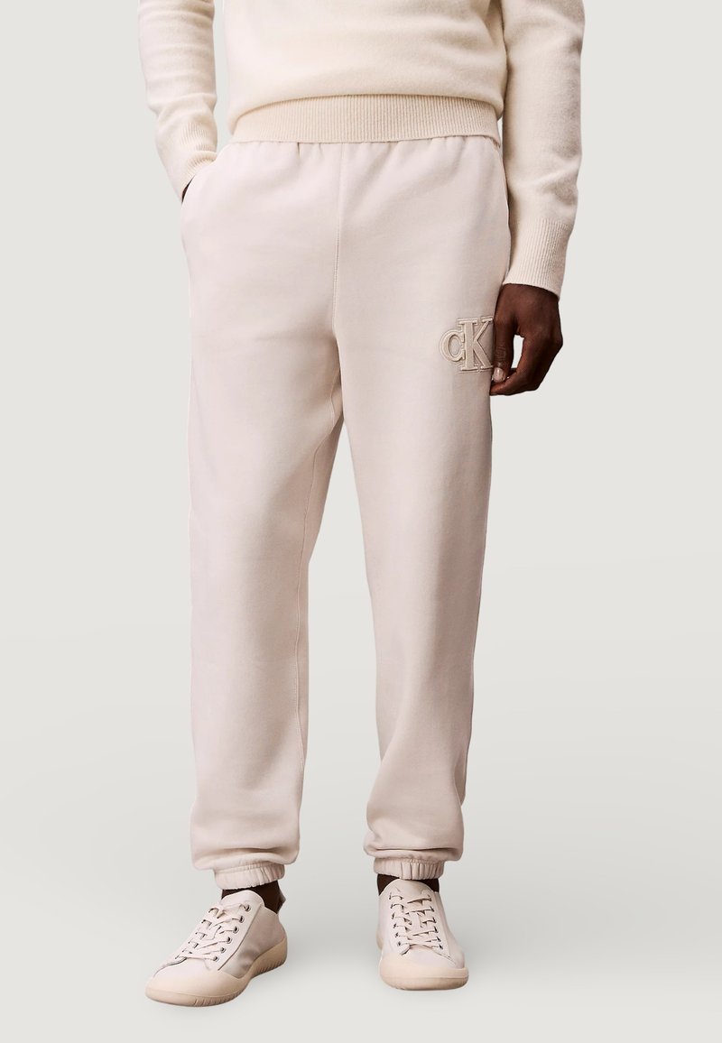 Beige katoenen joggers met een geribbelde tailleband, omgeslagen enkels en het geborduurde CK-logo aan de linkerkant, gecombineerd met bijpassende beige sneakers.