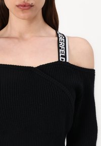 Schwarzer schulterfreier Pullover mit Rippstruktur, der ein überkreuztes Design und elastische Träger mit der Aufschrift "LAGERFELD" in Weiß und Grau aufweist.