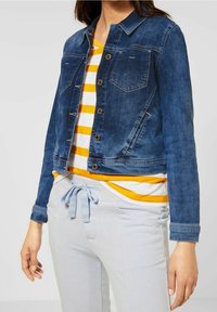 Džínsová bunda v tmavě modré barvě, s cropped designem, dvěma náprsními kapsami, zapínáním na knoflíky a jemným vyblednutím. Nositá přes tričko se žlutými pruhy.