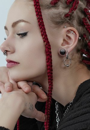 Braided red hair with silver and black jewelry. A septum ring, multiple earrings, and a visible tattoo on the hand resting under the chin.
