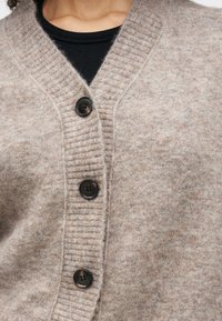 Šviesiai rudas kardiganas, pagamintas iš minkštos vilnos primenančios medžiagos, turintis griovelinį apykaklę, penkis juodus mygtukus ir struktūrinį paviršių.