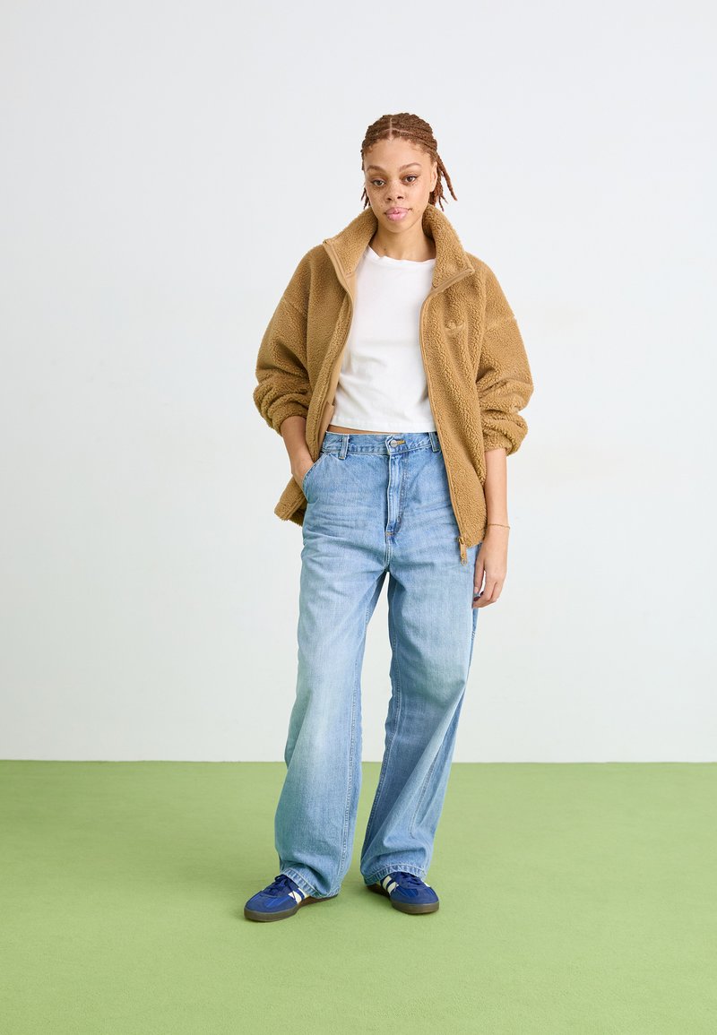 Bruine fleece zip-up jas, witte t-shirt, lichtblauwe oversized jeans en blauwe sneakers. Tegen een lichte achtergrond en een groene vloer.