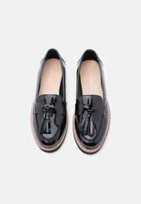 Melni lakoti mokasīni ar pušķu detaļu, apaļu purngalu un kontrastējošu beigu zoli. Gluda tekstūra ar minimālu šuvju skaitu.