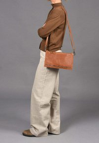 Bolso de piel de color marrón claro, tipo bandolera, con diseño plano, acentos de tachuelas plateadas en la parte frontal, correa ajustable y superficie lisa con textura.