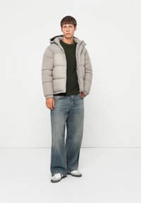 Lys grå polstret jakke med hette, glidelås foran og trykknapper. Mørkegrønn strikkegenser under. Løse blåjeans og joggesko.