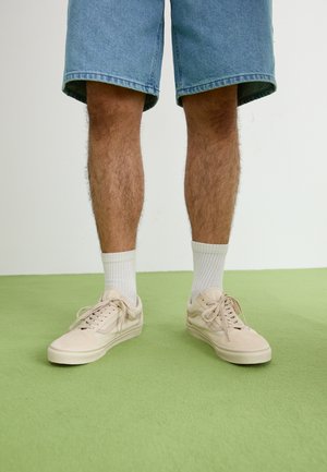 Jambes d’homme portant un short en jean bleu, des chaussettes blanches côtelées et des baskets beiges à lacets, debout sur un sol vert.