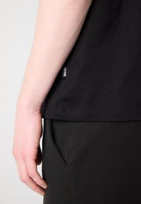 Gros plan de la main gauche d'une personne reposant près de la poche d'un pantalon noir, portant une chemise noire avec une étiquette "BOSS" visible.