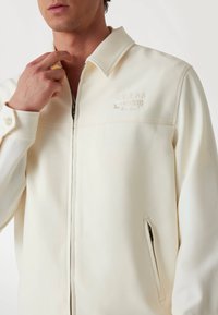 Light cream-colored jacket with a zip front, sturdy collar, and subtle logo embroidery on the chest. Features a side pocket.