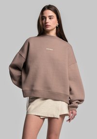 Pullover marrone oversize con finitura testurizzata, con un logo bianco sul petto e spalle scese, abbinato a una gonna beige chiara.