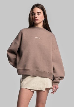 Oversized brown sweatshirt with a textured finish, featuring a white logo on the chest and dropped shoulders, paired with a light beige skirt.