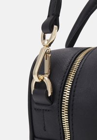 Gros plan sur le coin d'un sac en cuir noir montrant un fermoir en métal doré attachant une sangle et une fermeture éclair dorée le long du côté.