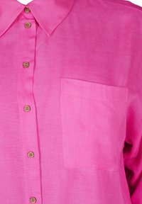 Košulja kratkih rukava, s gumbima, od svijetlo ružičastog lana. Ima ovratnik, pet drvenih gumba i džep na lijevoj strani prsiju. Glatka tekstura.