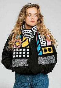 Kolorowy dziany szalik z geometrycznymi wzorami w kolorach czarnym, żółtym, różowym i turkusowym, z frędzlami na obu końcach.