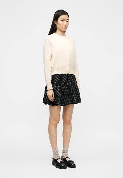 Νεαρή γυναίκα στέκεται φορώντας ανοιχτό μπεζ ζακέτα, μαύρη φούστα  με λευκές διαγώνιες ρίγες, μπεζ κάλτσες και μαύρα μοκασίνια σε λευκό φόντο.