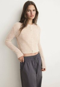 Hellrosa kurzes Strickjacke aus weichem Material mit langen Ärmeln und Knopfverschluss, kombiniert mit dunkelgrauen Hosen mit Kordelzug.