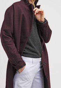 Burgundowa płaszcz z wysokim kołnierzem, z dużymi guzikami i luźnym krojem, noszony na szary sweter i jasne spodnie.