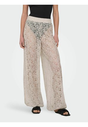 Femme portant un pantalon large en dentelle beige par-dessus des sous-vêtements noirs et des sandales noires, debout devant un fond clair uni.