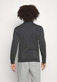 Tmavě šedý pulovr s dlouhým rukávem a vysokým límcem, s hladkým povrchem a žebrovanými manžetami. Nosený s světle šedými tepláky.