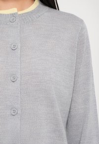 Cardigan grigio chiaro con scollatura rotonda, dotato di un colletto interno giallo e sei bottoni grigi lungo la parte anteriore. Texture morbida e lavorata a maglia.