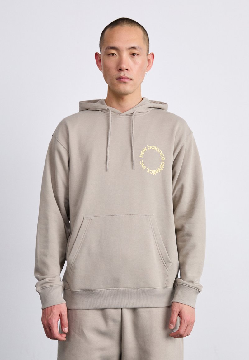 Hoodie beige en tissu doux avec une poche avant, capuche ajustable à cordon, et un logo circulaire jaune sur la poitrine gauche.