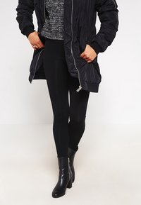 Svart vadderad bomberjacka med kviltat mönster, gråmönstrad tröja, svarta leggings och ankelskor. Enkel, funktionell outfit.