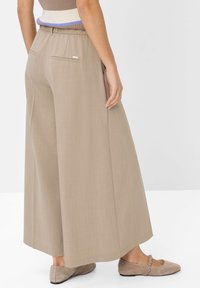 Donna che indossa pantaloni beige a gamba larga con tasche laterali e ballerine beige, in piedi contro uno sfondo bianco semplice.