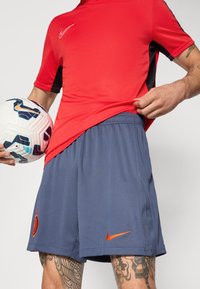Maillot de sport Nike rouge avec des accents noirs, associé à un short bleu marine arborant un logo Nike orange et un emblème d'équipe, tenu avec un ballon de soccer.