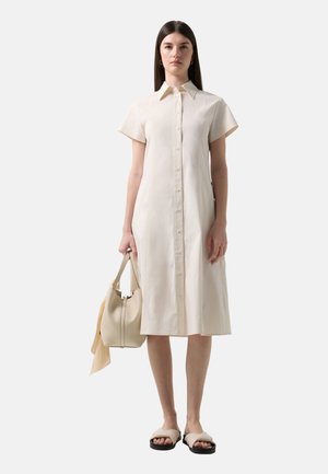 Kvinde iført en beige knælængde kjole med korte ærmer og knapper, der holder en beige taske og har beige sandaler på en hvid baggrund.