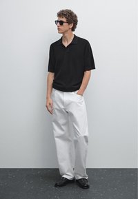 Polo shirt nero a maniche corte, pantaloni bianchi a gamba larga e scarpe nere. Occhiali da sole spessi e tessuto strutturato sono caratteristiche di design notevoli.