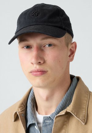 Ung mand med lys hud og blå øjne iført sort kasket, stribet skjorte, hvid undertrøje og beige jakke mod en ensfarvet lys baggrund.