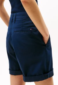 Granatowe szorty wykonane z gładkiego materiału, z podwiniętymi mankietami, dwiema tylnymi kieszeniami oraz małą metką z logo w czerwono-biało-niebieskich akcentach.