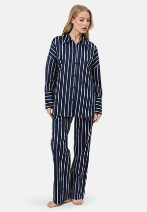 Kobieta w granatowej, długorękawowej koszuli na guziki i dopasowanych spodniach z białymi pionowymi paskami, stojąca boso na białym tle.