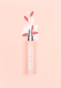 Rosa Glas-Sprühflasche mit silbernem Sprühkopf, beschriftet mit "IDAWARG LUXURIOUS FLOWER HAIR MIST", begleitet von weißen und roten Blütenblättern vor einem rosafarbenen Hintergrund.