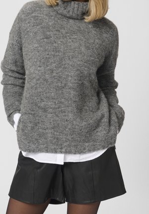 Femme portant un épais pull à col roulé gris sur une chemise blanche, associé à un short en cuir noir et des collants noirs transparents.