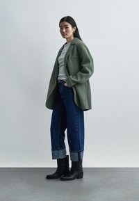 Γυναίκα όρθια, φοράει πράσινο φαρδύ σακάκι, γκρι πουλόβερ, σκούρα μπλε τζιν με γυρισμένα ρεβέρ, μαύρα μποτάκια αστραγάλου σε απλό γκρι φόντο.