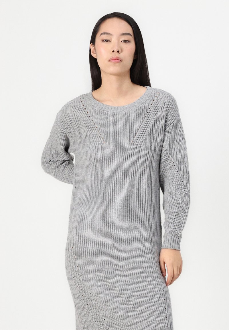 Długi szary sweter dzianinowy o ryflowanej fakturze, okrągłym dekolcie i subtelnych diagonalnych wzorach. Posiada długie rękawy i boczne rozcięcia.