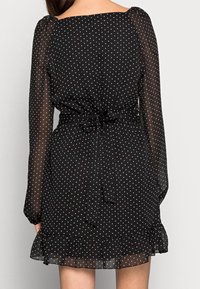 Černé šaty s bílými puntíky, vyrobené z průhledného materiálu, s dlouhými puff rukávy, vázáním v pase a volánkovým lemem, které mají malé bílé tečky po celém povrchu.