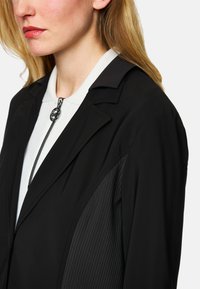 Schwarze Jacke mit einem taillierten Design, strukturierten Einsätzen und einem Reißverschluss mit einem runden Ziehgriff, getragen über einem hellen Oberteil.