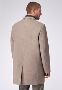 Beiger Wollmantel mit tailliertem Schnitt, ausgestattet mit einem Rückenschlitz, Kragen und dezent strukturierter Oberfläche. Nahtdetails an den Ärmeln.