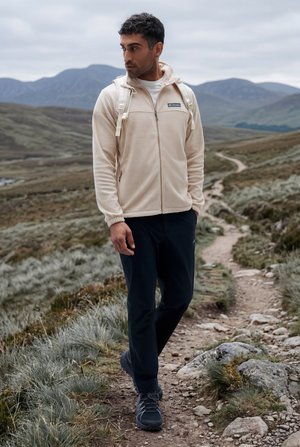 Vyras dėvi smėlio spalvos striukę ir kuprinę, žygiuojantis akmenuotu taku su kalnais fone po debesuotu dangumi.